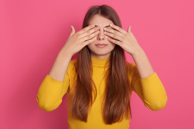 Horizontaal schot van vreedzame knappe leuke jonge model status geïsoleerd over roze muur in studio, die ogen behandelt met handen, geel sweatshirt draagt, die alleen is. Verrassing concept.