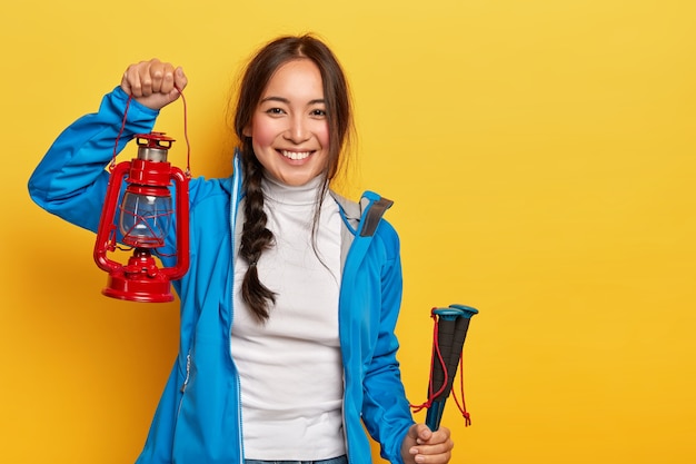 Horizontaal schot van vrolijke Aziatische vrouw met donkere vlecht, houdt gaslamp en wandelstokken, gekleed in actieve slijtage, heeft positieve glimlach staat over gele muur.