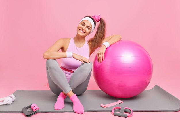 Horizontaal schot van vrolijke sportvrouw leunt op fitball doet gymnastiek thuis gekleed in sportkleding poses op fitnessmat die in goede fysieke vorm is geïsoleerd over roze achtergrond. Fitnesstraining