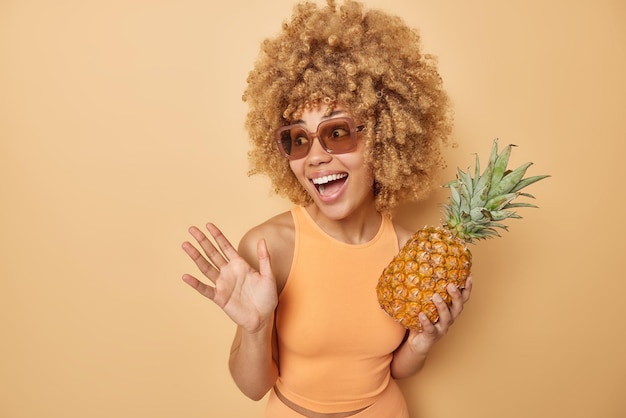 Horizontaal schot van vrolijke zorgeloze vrouw met krullend blond haar houdt verse ananas houdt palm omhoog lacht vrolijk draagt trendy zonnebril casual t-shirt geïsoleerd over beige achtergrond