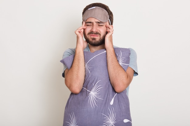 Horizontale bebaarde man poseert met gesloten ogen, houdt handen op zijn slapen, heeft vreselijke hoofdpijn, man draagt grijs t-shirt, knuffelt kussen, poseert met blinddoek op hoofd. Gezondheid concept.