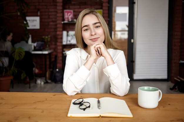 Horizontale opname van aantrekkelijke vriendelijke jonge vrouw in formele blouse, handen gevouwen en vreugdevol glimlachen naar de camera, gelukkig en geïnspireerd voelen tijdens het werken in café, notities maken, koffie drinken