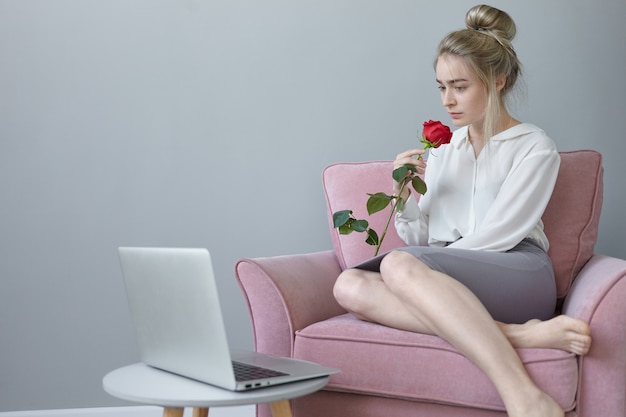 Horizontale opname van modieuze jonge vrouw in witte blouse en rok ontspannen in fauteuil, met een rode roos, zoete geur inademen, diep in gedachten, open laptop rustend op ronde tafel