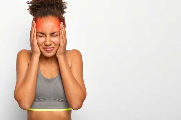 Horizontale opname van ontevreden gekrulde vrouw raakt beide slapen, lijdt aan migraine, draagt een sportbeha, sluit de ogen van slechte gevoelens, geïsoleerd op een witte muur