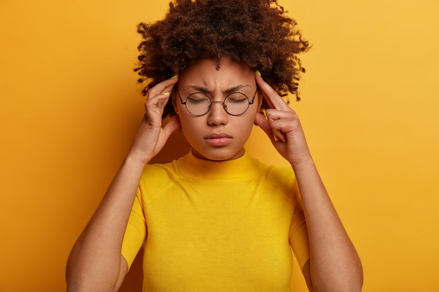 Gratis foto horizontale opname van ontevreden model met donkere huid voelt hoofdpijn, lijdt aan pijn in slapen, sluit ogen, heeft pijnstillers nodig, draagt een ronde bril en vrijetijdskleding, poseert tegen een gele muur