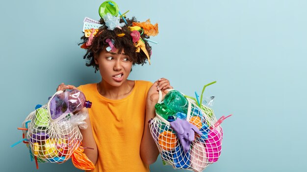 Horizontale opname van ontevredenheid Afro-Amerikaanse vrouw met donkere huid fronst gezicht als vuilnis oppikt, de omgeving reinigt, netzakken met plastic dingen vasthoudt, wegkijkt, vraagt om geen afval op de natuur te gooien