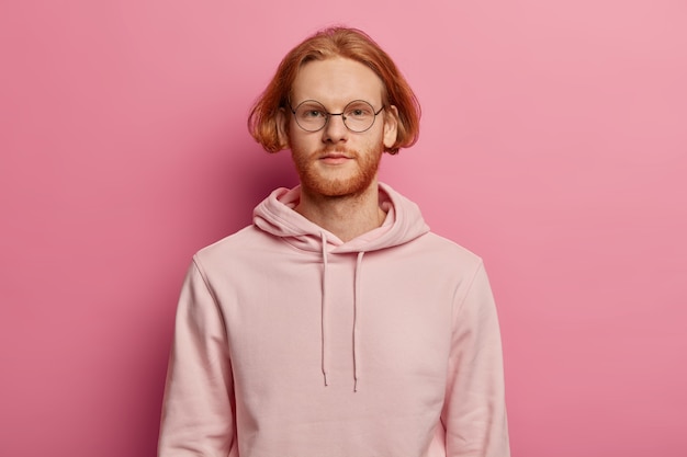 Horizontale opname van serieus uitziende bebaarde Europese man kijkt direct, heeft een rustige uitdrukking, draagt een casual hoodie, poseert tegen een roze muur, praat met iemand, heeft een bob-kapsel