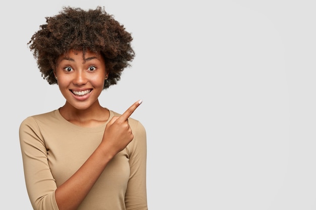 Horizontale opname van vrij donkere vrouw met Afro-kapsel, heeft een brede glimlach, witte tanden, toont iets aardigs aan vriend, wijst naar de rechterbovenhoek, staat tegen de muur