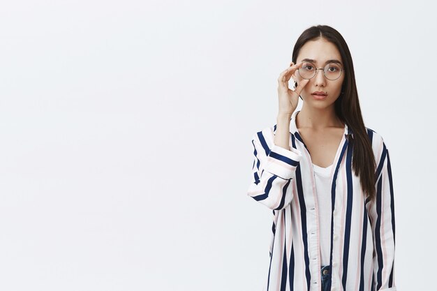 Horizontale schot van aantrekkelijke natuurlijke Aziatische vrouw met gebruinde huid en lang donker haar, ronde bril op de ogen aanraken, stijlvol gestreept shirt dragen over wit t-shirt, dromerig starend