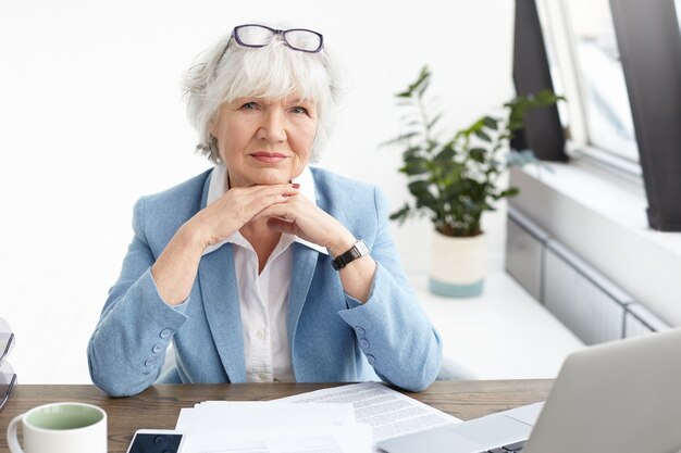 Horizontale schot van stijlvolle oudere vrouwelijke onroerend goed manager mooie blauwe pak en bril op haar hoofd dragen, handen onder de kin geklemd, met ernstige zelfverzekerde blik, laptop gebruikt voor werk