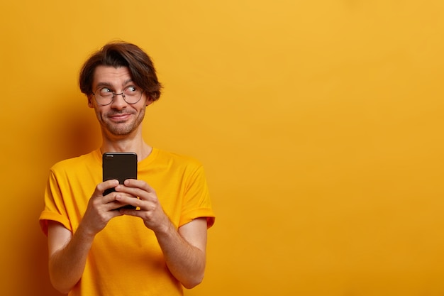 Horizontale schot van vrolijke hipster kerel houdt slimme telefoon en stuurt grappige foto's van feestje naar vriend, typen en bladert in netwerk, terloops gekleed, vormt tegen gele muur, kopie ruimte