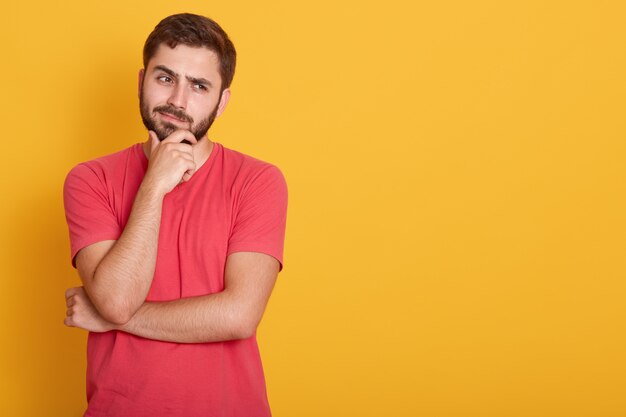 Horizontale serieuze ongeschoren man kleedt casual rood t-shirt, houdt hand onder kin, kijkt ernstig opzij, denkt aan iets, poseert op gele muur met vrije ruimte.