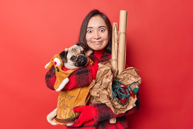 Horizontale shot jonge Aziatische vrouw poseert met favoriete hond draagt winter bovenkleding draagt opgerold papier en retro slinger voor vakantie decoratie geïsoleerd over levendige rode achtergrond Huisdieren concept