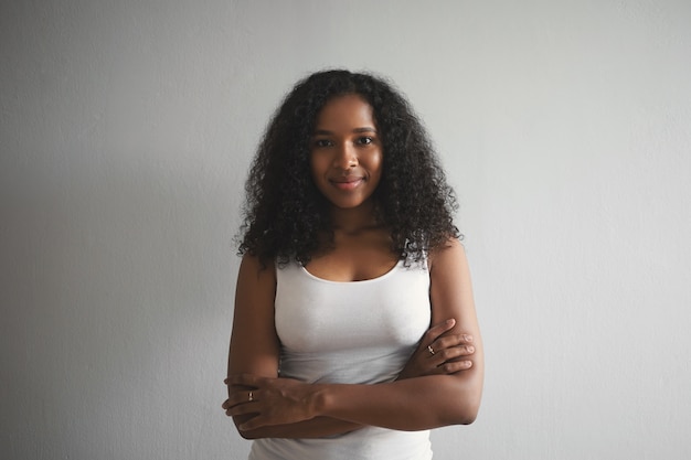 Horizontale stijlvolle mooie jonge Afro-Amerikaan in haar twintiger jaren poseren geïsoleerd binnenshuis met zelfverzekerde glimlach gekruiste armen op haar borst. Mensen, levensstijl en mode-concept