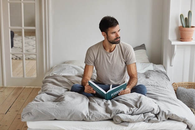 Horizontale weergave van doordachte bebaarde man houdt boek, zit gekruiste benen op comfortabel bed