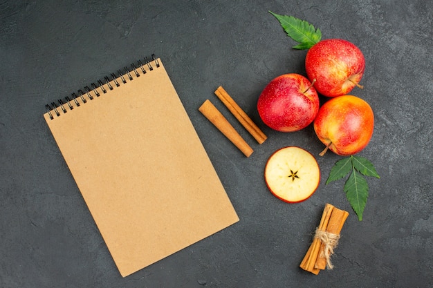 Gratis foto horizontale weergave van hele en gesneden verse natuurlijke biologische rode appels met groene bladeren, kaneellimoenen en notitieboekje op zwarte achtergrond