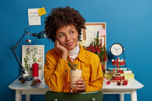 Horizontale weergave van peinzende vrouw gekleed in gele jas, advocaat cocktail houdt, geniet van heerlijke romige eimelk punch drinken