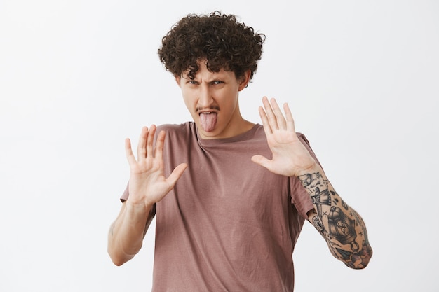 Houd dit ding bij mij vandaan. Stijlvolle getatoeëerde man die walging en afkeer uitdrukt en zijn handpalmen opheft om zichzelf te beschermen tegen voedsel, hij houdt niet van zijn tong uitsteekt en fronst tegen ontevreden emoties