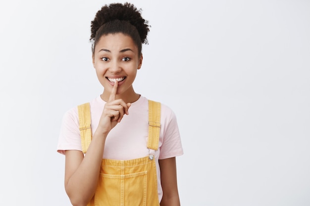 Houd geheimen veilig. Portret van charmante vriendelijk ogende schattige Afro-Amerikaanse vrouw in gele trendy overall, shh zeggen terwijl ze een stil gebaar toont, glimlachend en wijsvinger voor de mond houdt