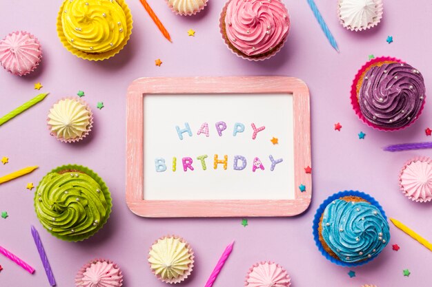 Houten gelukkige verjaardag op witte leisteen omringd met muffins; aalaw; kaarsen en hagelslag op roze achtergrond