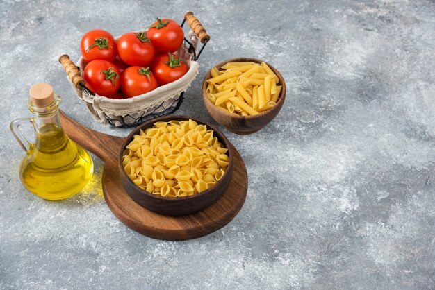 Houten kommen van verschillende rauwe pasta en verse tomaten op marmer.