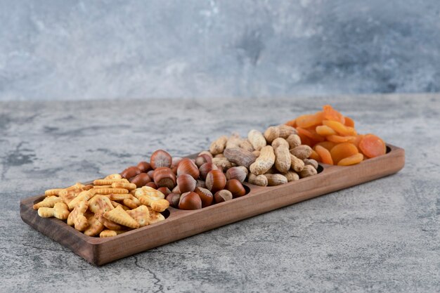 Houten plaat vol met verschillende noten, crackers en gedroogde abrikozen op marmeren ondergrond.