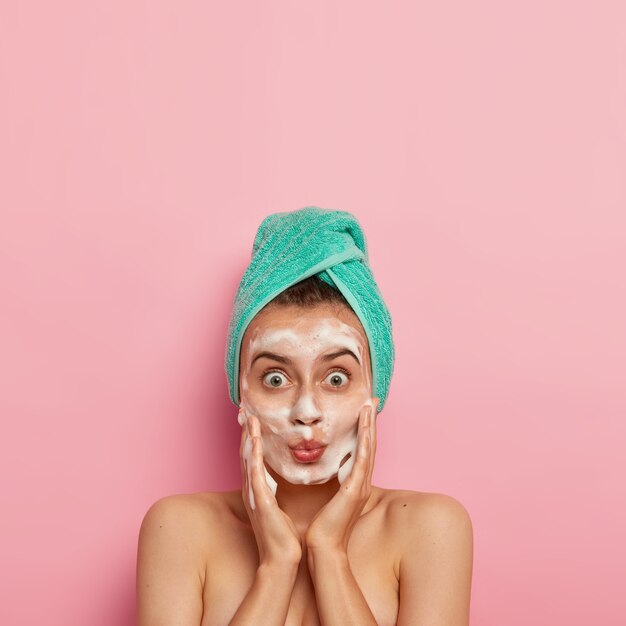 Huidverzorgingsroutine en hygiëneconcept. Foto van mooie vrouw met wijd geopende ogen, houdt de lippen gevouwen, masseert de wangen en brengt schuimende zeep aan