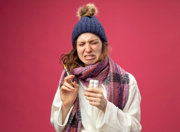 Huilend jong ziek meisje met witte mantel en winter hoed met sjaal met spuit met pillen geïsoleerd op roze