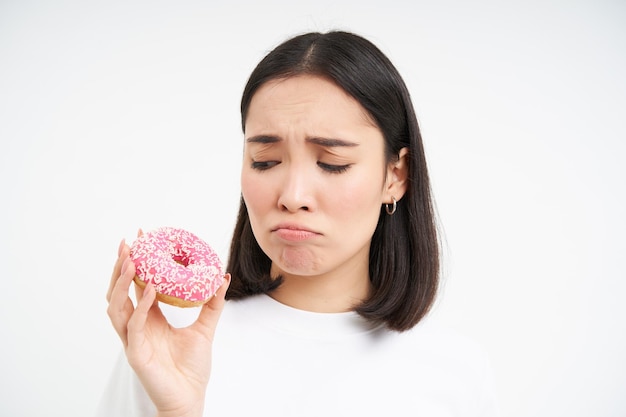 Gratis foto huilende aziatische vrouw die smakelijke douhnuts laat zien en snikkend op een gezond dieet is, kan geen junkfood wit eten