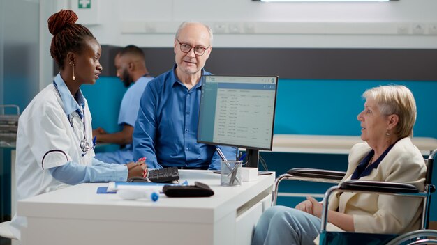 Huisarts praat met oude vrouw in rolstoel en haar man, doet controleconsult om te helpen bij herstelbehandeling. Medic die patiënt met chronische stoornis onderzoekt.