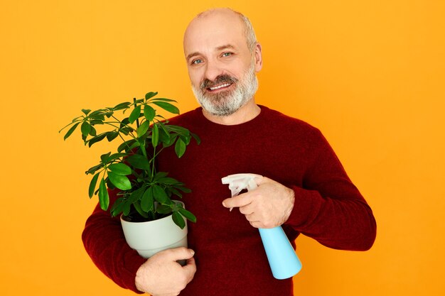 Huishouden, volwassen mensen, leeftijd en pensioen. Knappe emotionele bebaarde gepensioneerde man in trui helpen vrouw om huishoudelijk werk te doen beregening van water op groene plant met behulp van spuitfles, met vrolijke blik