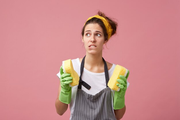 Huishouding, huishoudelijk werk, hygiëne en netheid. Gefrustreerde jonge vrouw in schort en beschermende handschoenen die op haar lip bijten, zich gestrest voelen omdat ze de kamers niet haalt voordat de gasten komen