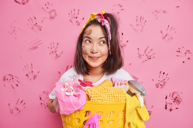 huisvrouw met donker haar poseert in de buurt van een mand vol vuile was heeft een vies gezicht geïsoleerd op roze