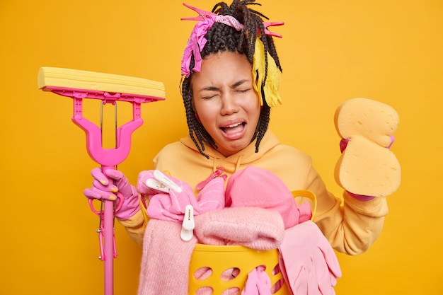 huisvrouw moe van het schoonmaken van het huis de hele dag houdt spons en dweil vast en drukt negatieve emoties uit, gekleed in casual huishoudelijke kleding geïsoleerd op geel