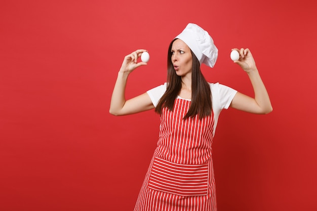 Huisvrouw vrouwelijke chef-kok of bakker in gestreepte schort, wit t-shirt, toque chef-koks hoed geïsoleerd op rode muur achtergrond. leuke huishoudster vrouw houdt in handen twee kippeneieren. mock-up kopie ruimteconcept.
