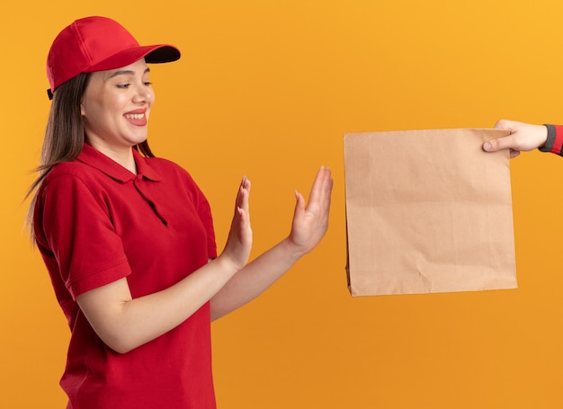 Iemand geeft een papieren pakket aan een tevreden mooie bezorger in uniform, hand in hand open gebaren zonder teken geïsoleerd op oranje muur met kopie ruimte