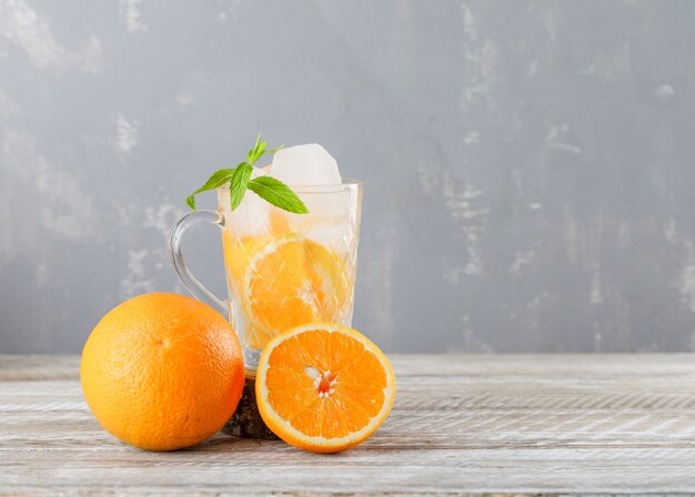 IJzig detoxwater in een kop met sinaasappelen, munt zijaanzicht over houten en pleisterachtergrond