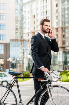 Ik ben zo. zelfverzekerde jonge zakenman die zijn fiets rolt en op de mobiele telefoon praat terwijl hij buiten loopt