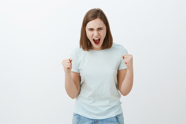 Ik haat regels. Portret van verontwaardigde en pissige boze jonge vrouw met kort bruin haar, kijkend van onder het voorhoofd, fronsend en mokkend, gebalde vuisten van woede en boosheid die geduld verliest over grijze muur