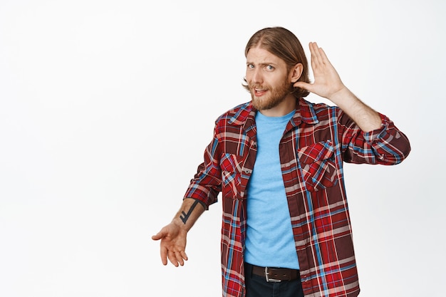 Ik kan je niet horen. Verwarde blonde man houdt de hand bij het oor en kijkt verbaasd, vertelt luider te spreken, kan niet begrijpen wat de persoon zegt, staande op wit.