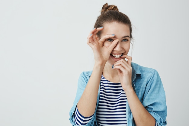 Ik richt op jou. Portret van speelse gelukkig goed uitziende vriendin met knot kapsel ok teken over oog houden en op zoek dacht het, lip met de hand aanraken en breed glimlachend