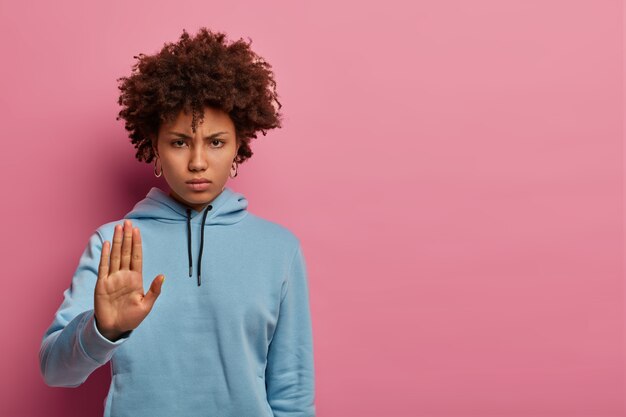 Ik weiger. Boze nors vrouw met krullend haar steekt handpalm op of verbodsgebaar, zegt nee, wacht even, wacht even, grijnzend gezicht, draagt blauwe hoodie, modellen over roze pastelkleurige muur, kopieer ruimte opzij.