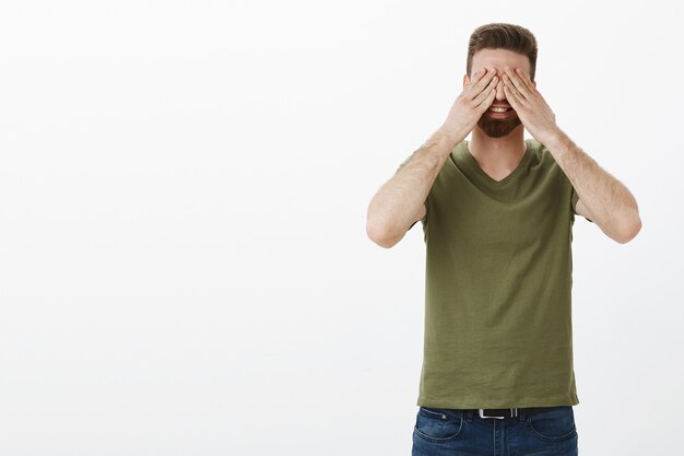 Ik zweer het niet te kijken. Portret van knappe charmante blanke man met baard sluit ogen met palmen in anticiaption, glimlachend vreugdevol wachtend op verrassing