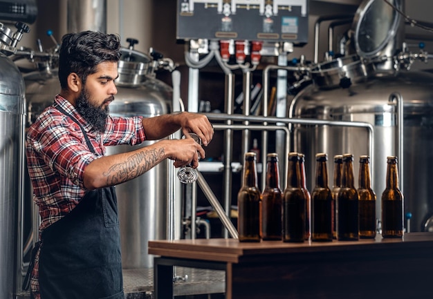 Indiase bebaarde getatoeëerde hipster mannelijke fabrikant die ambachtelijk bier presenteert in de microbrouwerij.