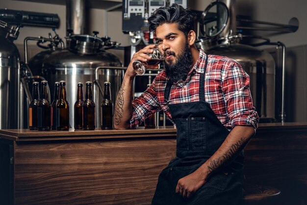 Indiase bebaarde getatoeëerde hipster mannelijke fabrikant die ambachtelijk bier proeft in de microbrouwerij.