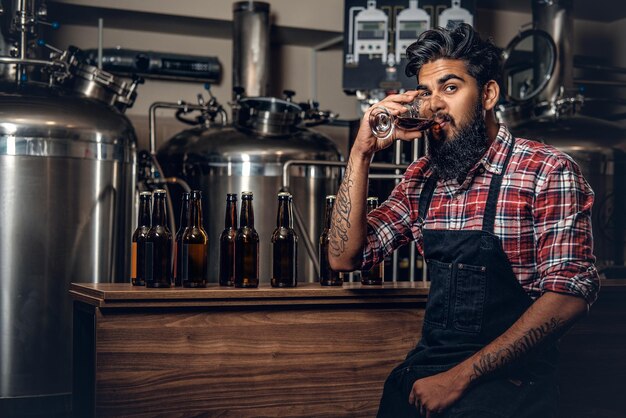 Indiase bebaarde getatoeëerde hipster mannelijke fabrikant die ambachtelijk bier proeft in de microbrouwerij.