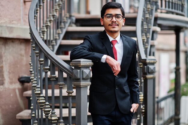 Indiase jongeman met bril draagt een zwart pak met rode stropdas buiten tegen een ijzeren trap