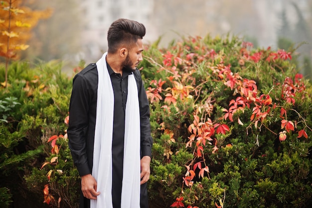 Indiase stijlvolle man in zwarte traditionele kleding met witte sjaal buiten geposeerd tegen gele herfstbladeren boom