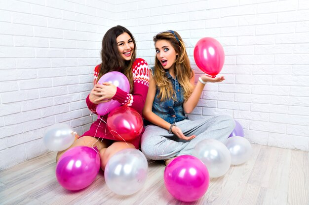 Indoor helder portret van twee grappige weddenschap vrienden zus hipster dames, gek, feest, roze ballon, v wetenschap, knuffels en kusjes, make-up, truien, geweldige glimlach tonen.