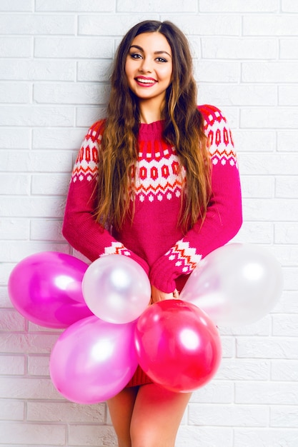 Indoor lifestyle beeld van grappige mooie brunette meisje met lichte make-up en lange haren, trendy trui dragen en roze partij ballonnen te houden.